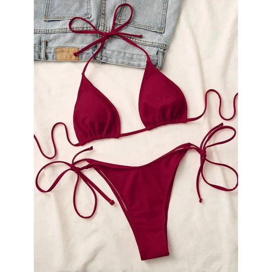 Bikini triangle bordeaux à lacets de nouage sur les côtés, posé à plat sur un tissu blanc avec un short en jean