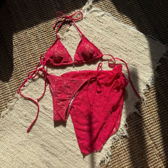 Maillot de bain trois pièces avec paréo rose fuchsia en dentelle posé à plat sur une natte, paréo noué sur le côté
