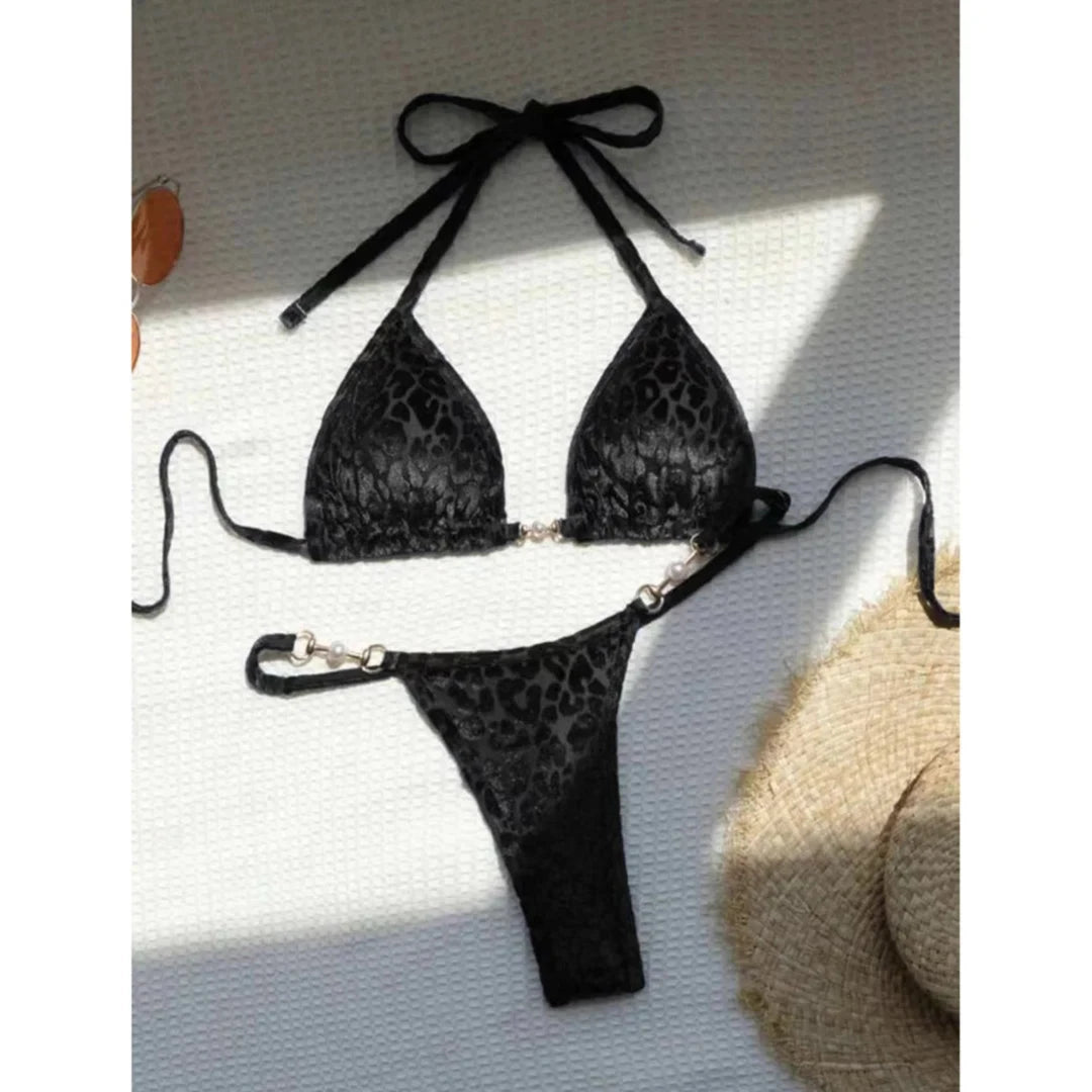 Bikini string noir imprimé léopard posé à plat sur un tissu gris, haut triangle à lien et bas à anneaux métalliques