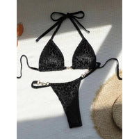 Bikini string noir imprimé léopard posé à plat sur un tissu gris, haut triangle à lien et bas à anneaux métalliques
