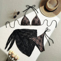 Maillot de bain trois pièces avec paréo noir en dentelle posé à plat sur un tapis, lunettes et chapeau de paille