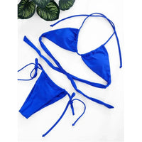 Bikini string mini tour de cou taille basse bleu roi posé à plat sur fond blanc, feuille tropicale en haut à gauche