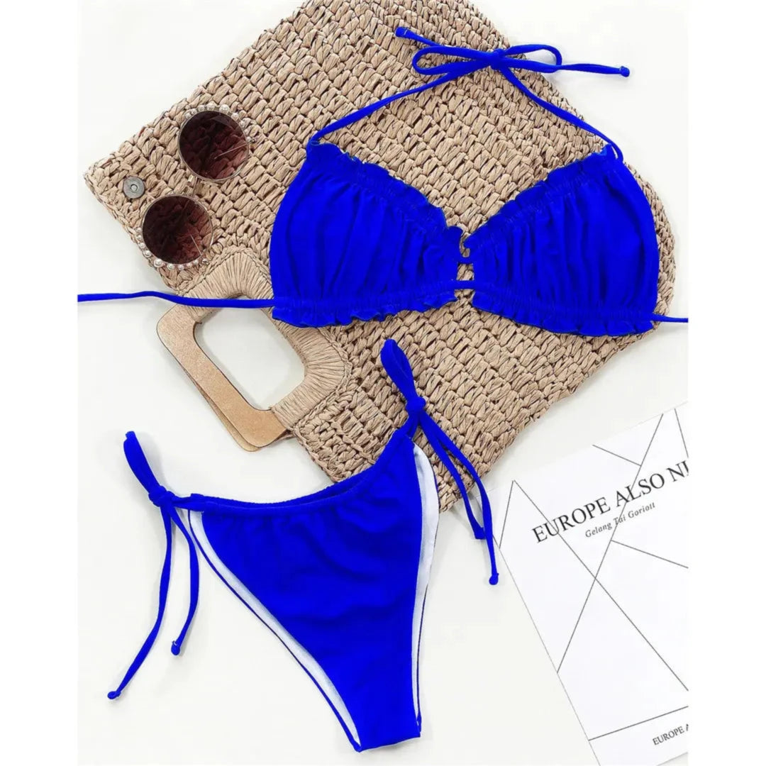 Bikini à volants froncé col halter bas à nouer bleu roi à plat sur un sac en paille, à côté d'un magazine