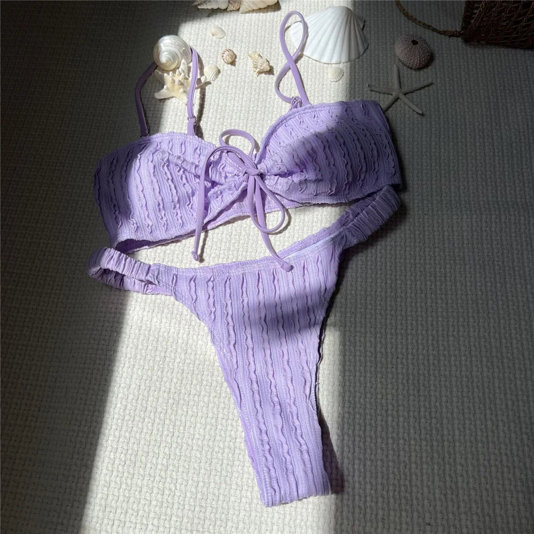Bikini brésilien texturé froissé à nœuds violet posé à plat sur un tapis, ombre du soleil et coquillages autour