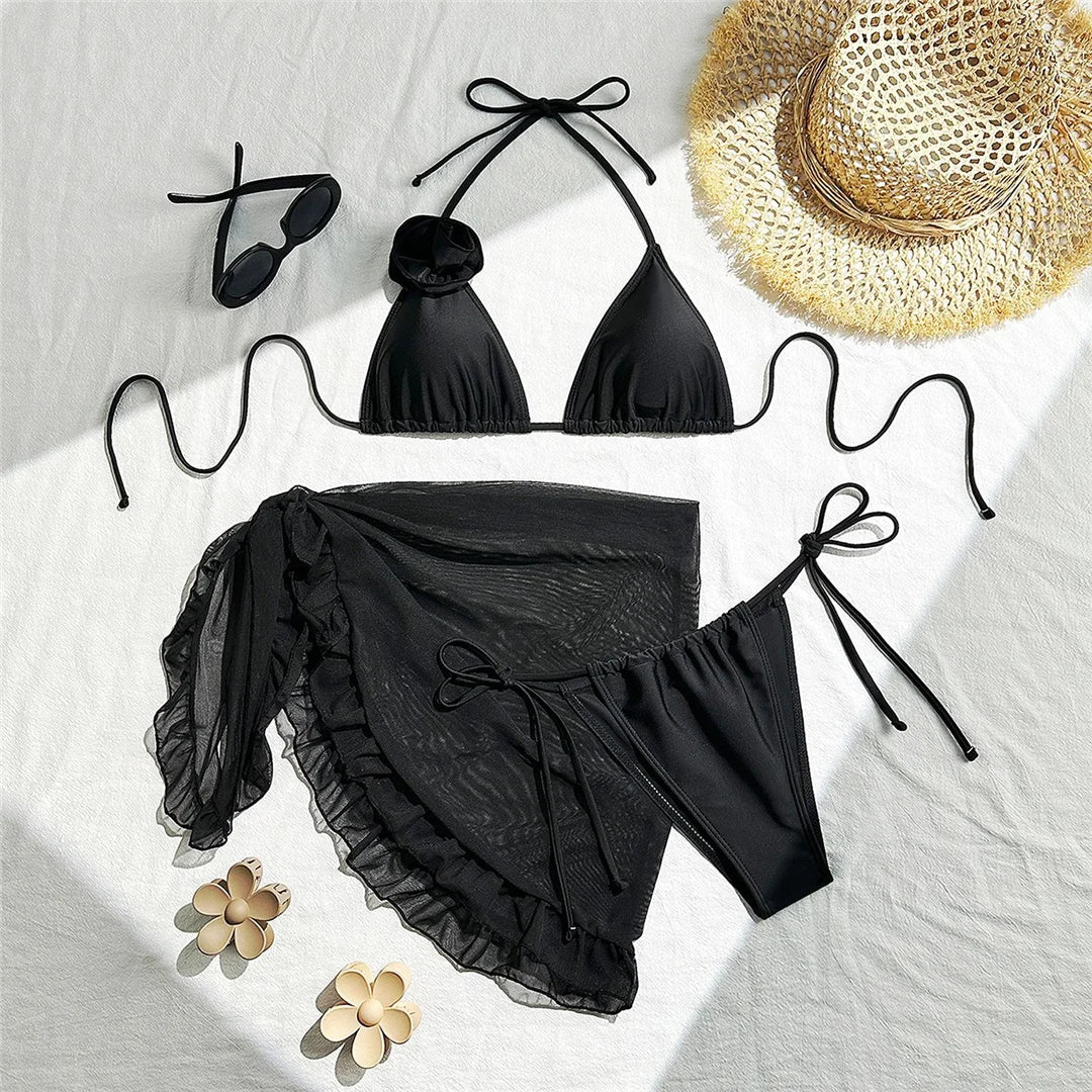 Maillot trois pièces noir à plat sur un drap blanc, avec pareo transparent à volants, chapeau de paille et lunettes de soleil