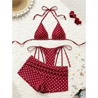Maillot trois pièces à pois rouge posé à plat sur une surface blanche, avec lunettes et petites fleurs