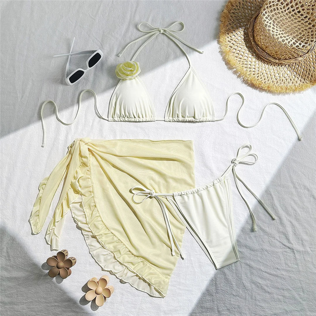 Maillot trois pièces blanc cassé à plat sur un drap blanc, avec pareo à volants, lunettes de soleil et chapeau de paille