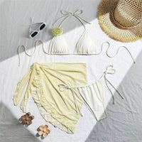 Maillot trois pièces blanc cassé à plat sur un drap blanc, avec pareo à volants, lunettes de soleil et chapeau de paille