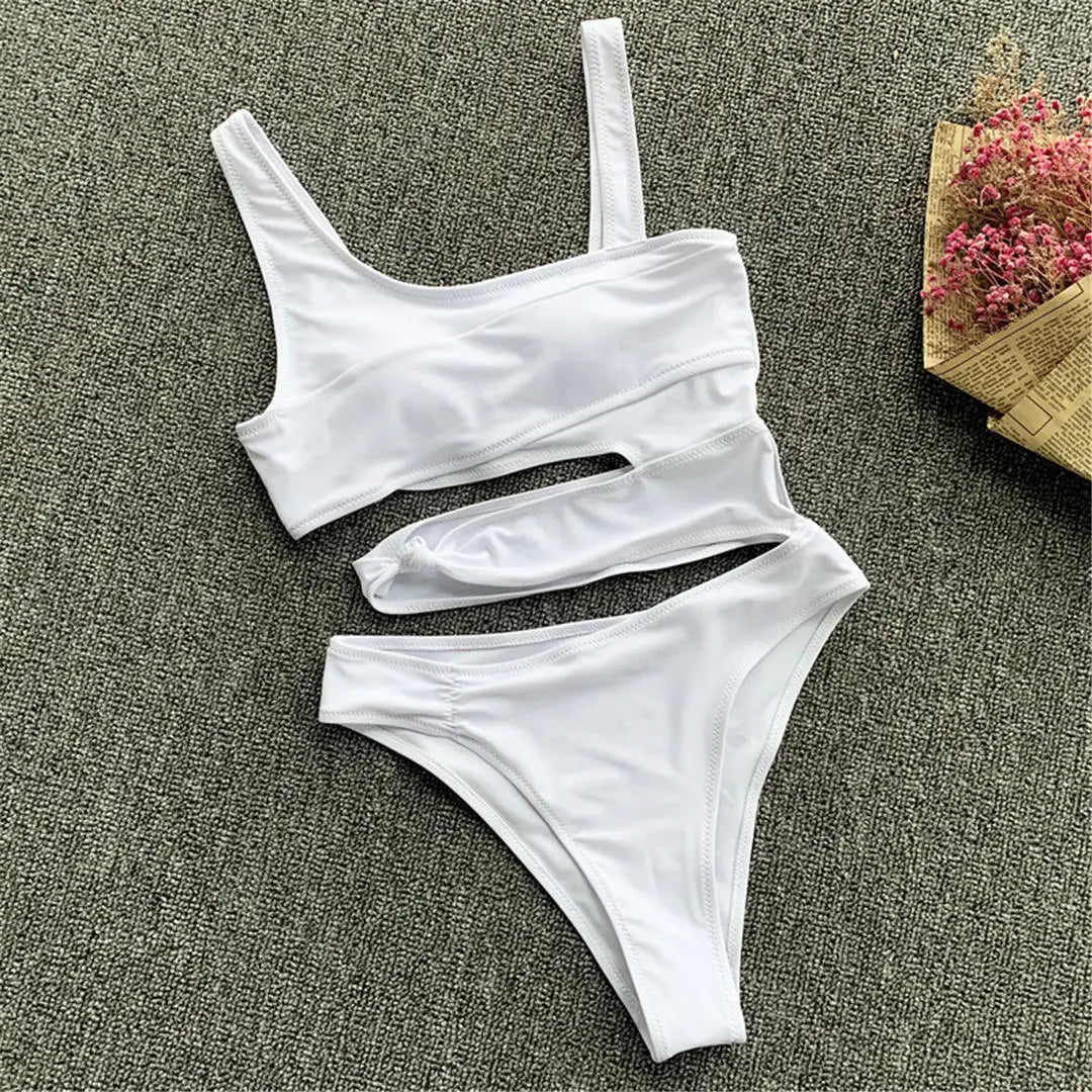 Maillot de bain une pièce échancré blanc posé à plat sur un tapis gris, bretelle unique et découpes horizontales