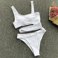 Maillot de bain une pièce échancré blanc posé à plat sur un tapis gris, bretelle unique et découpes horizontales