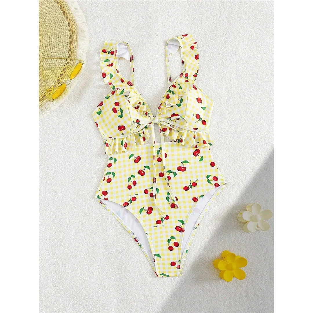 Maillot de bain une pièce à volants échancré jaune clair à cerises posé à plat, à côté d'un chapeau de paille
