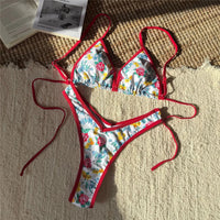 Bikini brésilien string taille basse imprimé fleuri rouge posé à plat sur un tapis, liseré rouge et liens latéraux longs