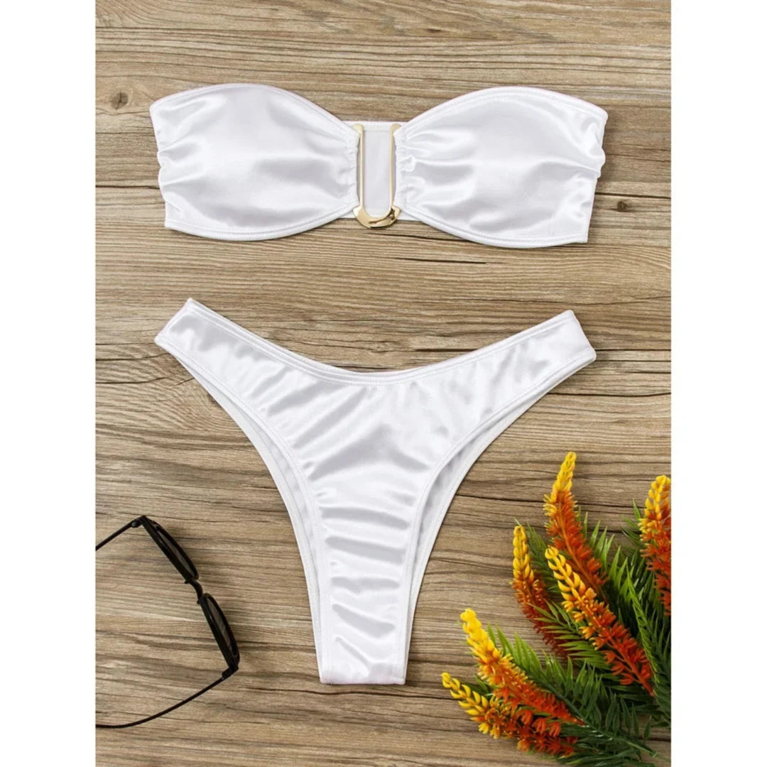Bikini bandeau sans armatures taille basse blanc posé à plat sur bois clair, lunettes de soleil et fleurs à côté