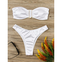 Bikini bandeau sans armatures taille basse blanc posé à plat sur bois clair, lunettes de soleil et fleurs à côté