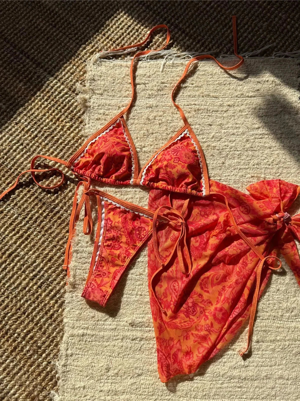 Maillot trois pièces rouge orangé posé à plat sur un tapis en osier, haut triangle à liseré blanc et slip ficelé