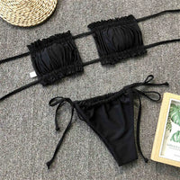 Bikini bandeau à volants bretelles réglables noir posé à plat sur un tapis gris, à côté d'un magazine