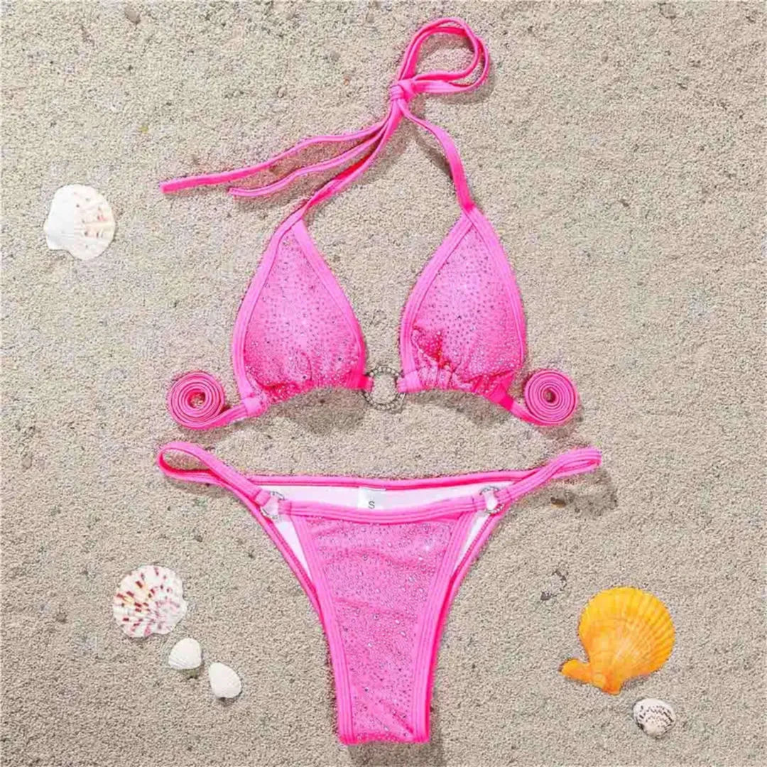 Bikini mini strass sans armatures taille basse rose posé à plat sur du sable, strass et anneaux sur le haut