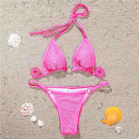 Bikini mini strass sans armatures taille basse rose posé à plat sur du sable, strass et anneaux sur le haut