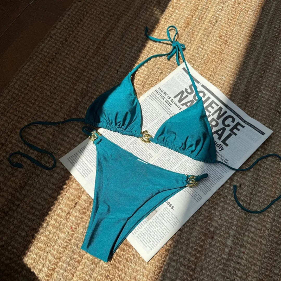Bikini tour de cou à anneau métallique bleu paon posé à plat sur un tapis en fibres, haut triangle et bas aux anneaux dorés