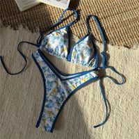 Bikini brésilien string taille basse imprimé fleuri bleu posé à plat sur un tapis, liseré bleu et liens latéraux longs