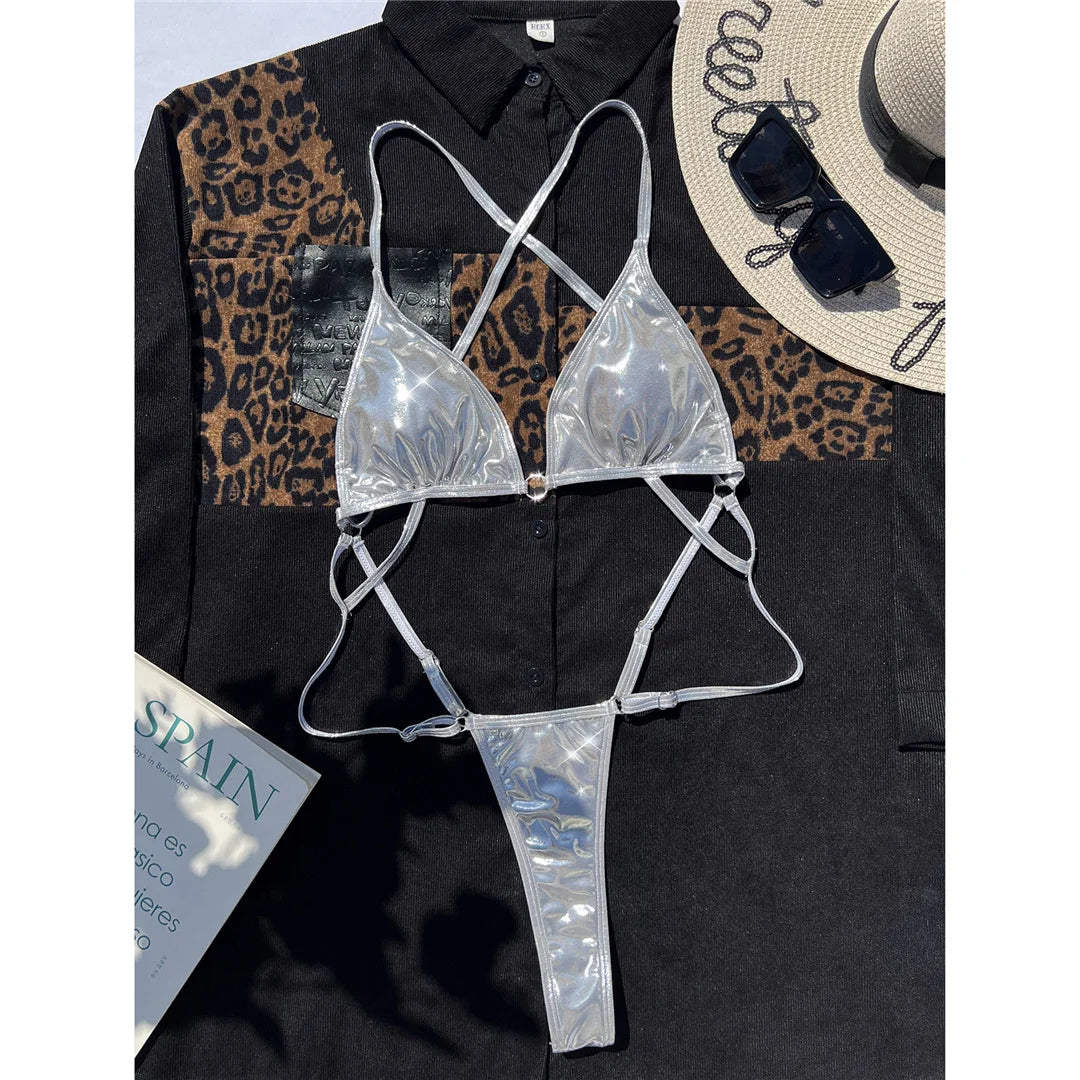Maillot une pièce string argenté effet simili cuir posé à plat sur une chemise noire, avec un chapeau de paille et un magazine