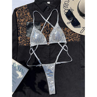 Maillot une pièce string argenté effet simili cuir posé à plat sur une chemise noire, avec un chapeau de paille et un magazine