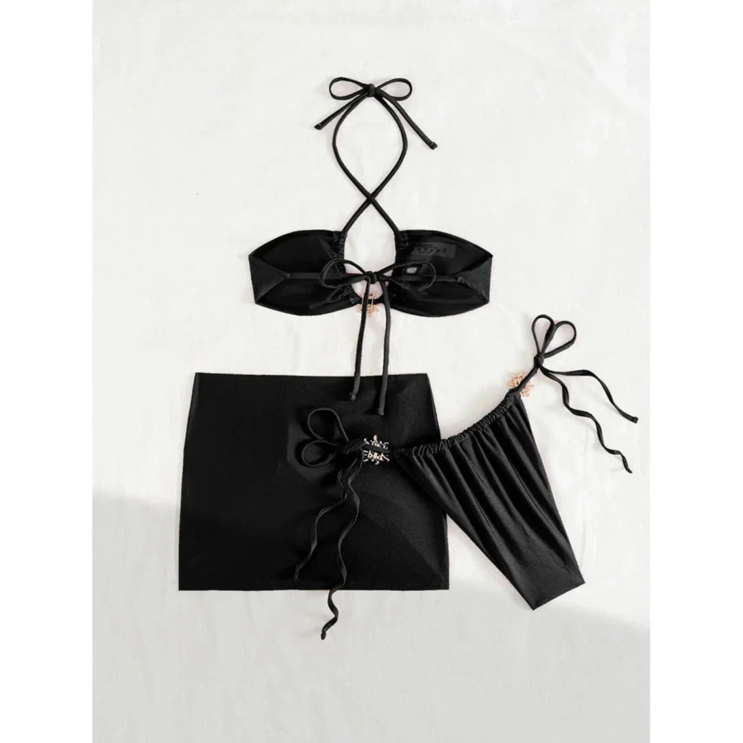 Maillot de bain trois pièces jupe halter à brides noir photographié à plat sans accessoires, nœud central sur la jupe et bas latéral