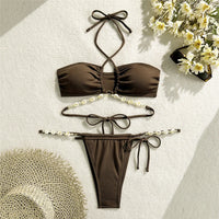 Bikini marron à coquillages posé à plat sur un tapis de sable, avec un chapeau de paille et des fleurs blanches