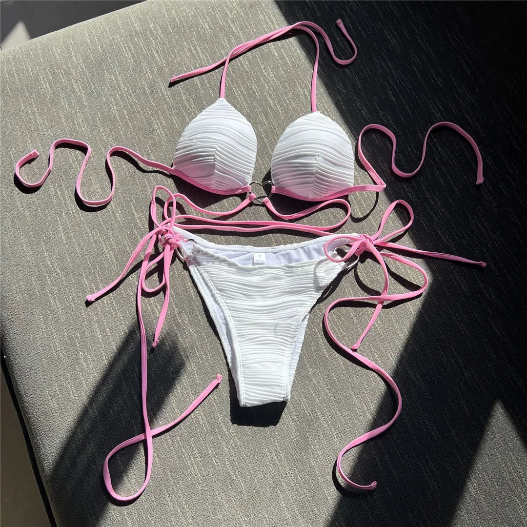 Bikini col halter croisé à effet froissé blanc posé à plat sur un coussin gris, longs lacets roses et slip à nouer