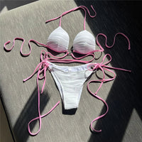 Bikini col halter croisé à effet froissé blanc posé à plat sur un coussin gris, longs lacets roses et slip à nouer