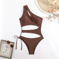 Maillot une pièce marron à découpes posé à plat, bretelle asymétrique et lien noué sur le côté, près d'un chapeau