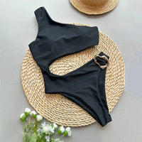 Maillot de bain une pièce asymétrique à anneau métal noir posé à plat sur un set en osier avec fleurs blanches