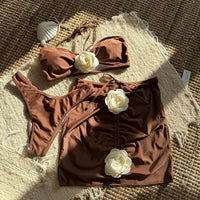 Maillot de bain trois pièces jupe fleurs en relief marron café à plat sur un tapis, avec coquillages et fleurs blanches