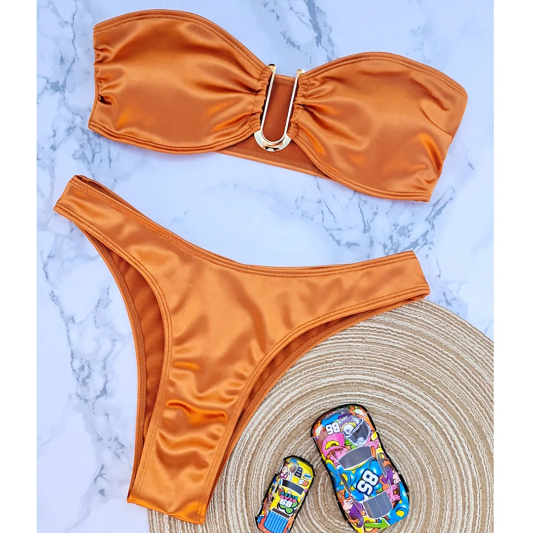 Bikini bandeau sans armatures taille basse orange posé à plat sur marbre blanc, tapis et objets à côté, vue de dessus