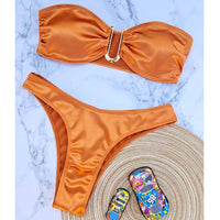 Bikini bandeau sans armatures taille basse orange posé à plat sur marbre blanc, tapis et objets à côté, vue de dessus
