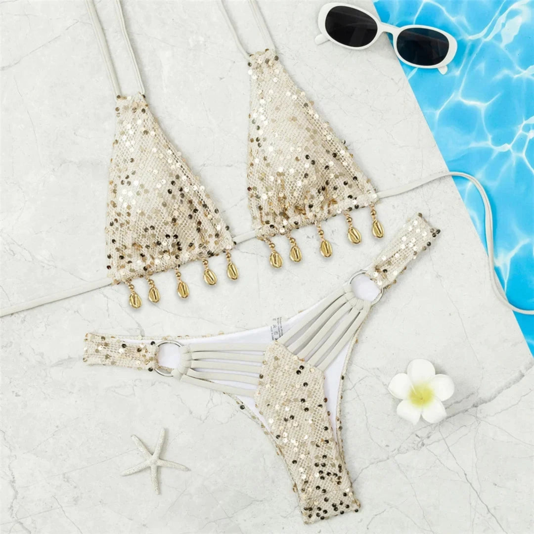 Bikini brésilien à sequins tour de cou beige posé à plat sur du marbre, haut à franges de coquillages et bas pailleté
