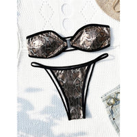 Bikini bandeau échancré imprimé peau de serpent posé à plat sur une étoffe blanche, bordures noires et string fin