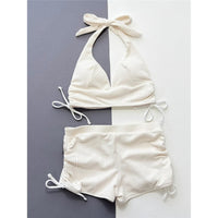 Bikini taille haute beige côtelé posé à plat sur fond gris et blanc, haut à col halter et short à liens latéraux