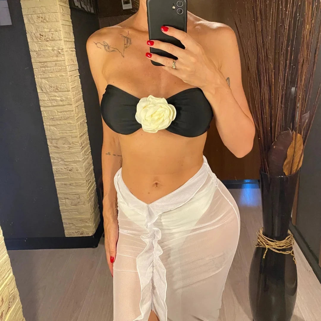 Maillot trois pièces bandeau porté de face par une femme, rose en relief blanche sur bandeau noir et paréo blanc en mesh noué devant
