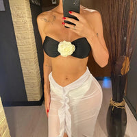 Maillot trois pièces bandeau porté de face par une femme, rose en relief blanche sur bandeau noir et paréo blanc en mesh noué devant