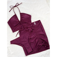 Maillot trois pièces avec jupe rouge bordeaux posé à plat sur un tissu blanc, haut à lacets, slip et jupe froncée