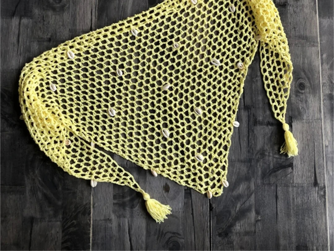 Paréo en crochet ajouré jaune à coquillages posé à plat sur bois sombre, avec fleurs blanches en haut à droite
