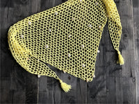 Paréo en crochet ajouré jaune à coquillages posé à plat sur bois sombre, avec fleurs blanches en haut à droite