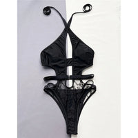 Maillot une pièce noir à col tour de cou, découpe en V au décolleté et partie basse en dentelle, posé à plat sur fond gris et blanc