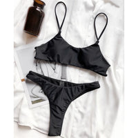 Bikini string brassière noir posé à plat sur une chemise blanche, à côté d'une bouteille brune et d'un magazine