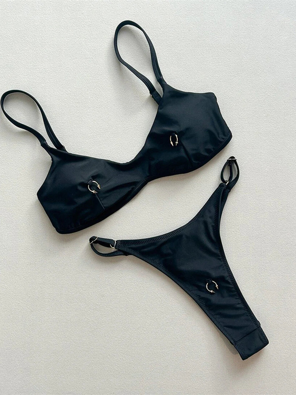 Bikini string micro triangle à anneaux métalliques noir posé à plat sur fond clair, anneaux ronds sur le haut et le bas