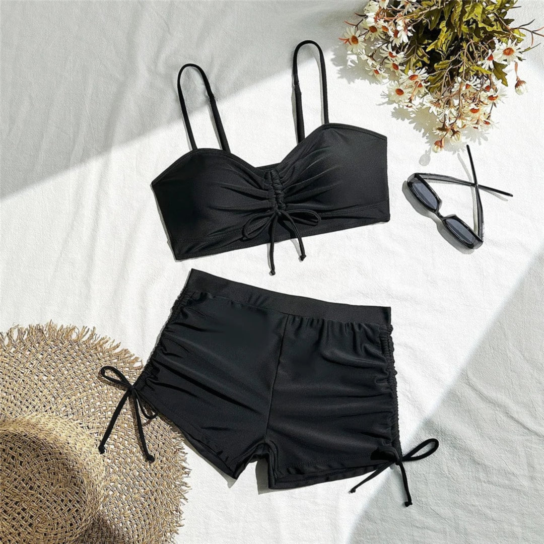 Bikini taille haute short froissé noir étalé à plat sur un drap blanc, avec un chapeau de paille, des lunettes et des fleurs
