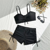 Bikini taille haute short froissé noir étalé à plat sur un drap blanc, avec un chapeau de paille, des lunettes et des fleurs