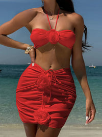 Maillot de bain trois pièces jupe fleurs en relief rouge porté de face sur la plage, main sur la hanche, bustier à fleur