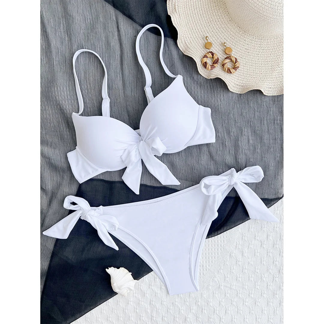 Bikini push-up noué avec armatures et rembourrage blanc posé à plat sur tissu gris, nœud central et culotte nouée sur le côté